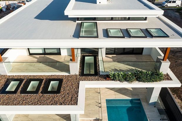 Aerial view of a modern flat-roof residential building with extensive rooftop glazing, white rendered parapets, pale tiled terraces, landscaped gravel roof areas, and a small rectangular swimming pool. The composition highlights a geometric, low-profile architectural form with abundant skylights and outdoor courtyards.