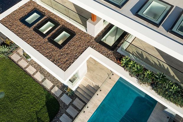 Aerial view of a contemporary flat-roofed residence with extensive rooftop gravel areas, multiple skylights, large glazed openings, a paved pool terrace, rectangular swimming pool, landscaped planting, and stepping-stone paths. The building uses crisp white roof/parapet edges and dark-framed glazing.