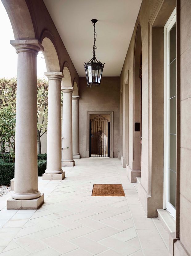 A covered residential entry colonnade with a long tiled walkway, repeated classical columns and rounded arches, stucco walls, a hanging lantern, and a decorative metal entry gate at the far end. A glazed entry door is visible on the right, with landscaped greenery beyond the open side of the veranda.