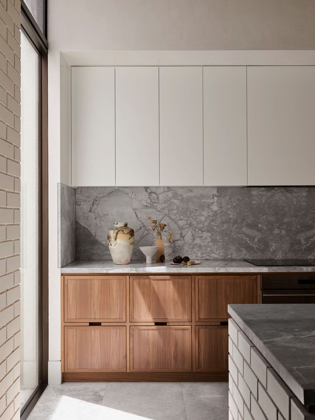 A refined contemporary kitchen with a compact galley-style layout. The space features floor-to-ceiling white cabinetry, richly grained timber lower cabinets, and a dramatic grey veined stone splashback and benchtop. A stone-clad island or peninsula occupies the foreground, while a tall window and light brick wall bring natural light into the room. Minimal decorative objects sit on the counter.