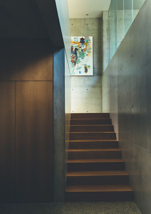 A narrow, modern stairway with dark timber treads rising between raw concrete walls. A dark timber-paneled built-in surface lines the left side, while a clear glass balustrade protects the upper landing. A colourful artwork is mounted on the concrete wall at the top of the stairs, and the lower floor has a speckled polished finish.