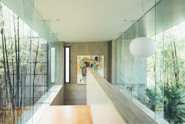 A bright contemporary interior corridor or landing with extensive floor-to-ceiling glazing on both sides, opening views to dense bamboo and tropical planting. A long pale concrete or stone-clad central partition leads toward a colourful artwork on a textured concrete wall. The space has a smooth white ceiling with recessed downlights, minimalist detailing, a large spherical pendant light, and warm timber flooring or a timber surface in the foreground.