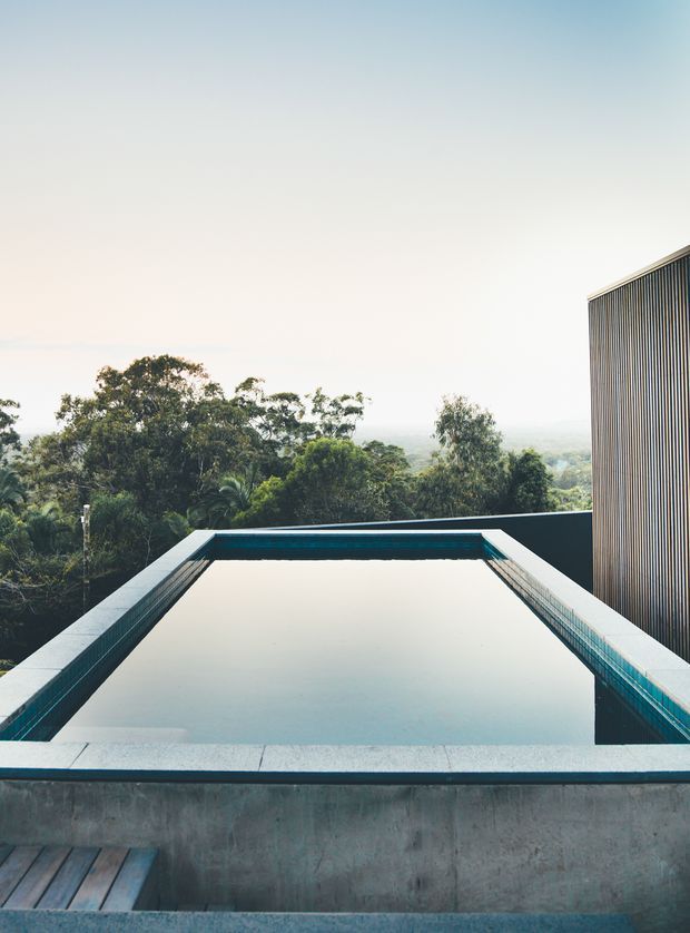 A contemporary rooftop or elevated infinity-edge swimming pool viewed from the deck, with dark tiled pool surrounds, exposed concrete walls, and a vertical slatted building facade at right. Lush treetops and a hazy distant landscape extend beyond the pool, creating a serene elevated outlook.