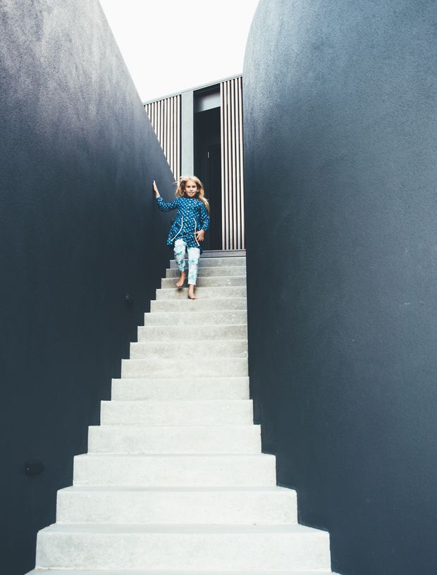 A narrow exterior stairway rises between two tall dark grey textured walls. The broad concrete steps lead to a black entry door framed by vertical timber battens, with a child descending the stairs. The composition emphasizes the dramatic enclosed architectural passage and strong contrast between pale concrete and dark walls.
