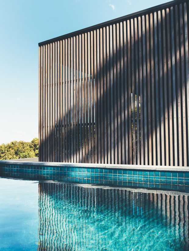 Exterior architectural detail showing a swimming pool beside a tall screened facade made of closely spaced vertical dark slats. The slatted wall creates strong linear shadows and reflections in the turquoise pool water, with clear sky and distant vegetation in the background.