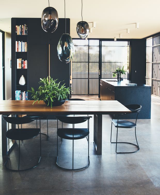 A contemporary open-plan dining and kitchen area with a long timber dining table, black metal-framed dining chairs, sculptural smoked-glass pendant lights, dark built-in shelving, indoor plants, and a compact dark kitchen island. Full-height black vertical screens and glazing create a bright, layered backdrop. The polished concrete floor and restrained black, timber, and neutral palette give the space a refined industrial-modern character.