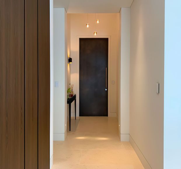 A narrow, refined residential hallway or entry passage viewed toward a dark timber or painted feature door. The space has pale walls, light stone or tile flooring, slim white skirting and door surrounds, a tall timber built-in surface at left, a small console table, and three warm pendant lights creating a softly illuminated, contemporary interior.