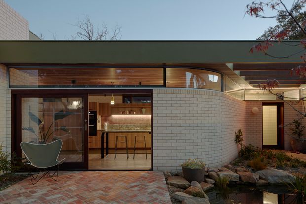 Exterior view of a low-slung contemporary residence at dusk, with a broad flat roof, dark-framed glazing, warm timber-lined clerestory windows, and a curved white brick wall enclosing an illuminated kitchen and entry. A brick-paved courtyard with rocks, planting, and a small reflective water feature occupies the foreground.