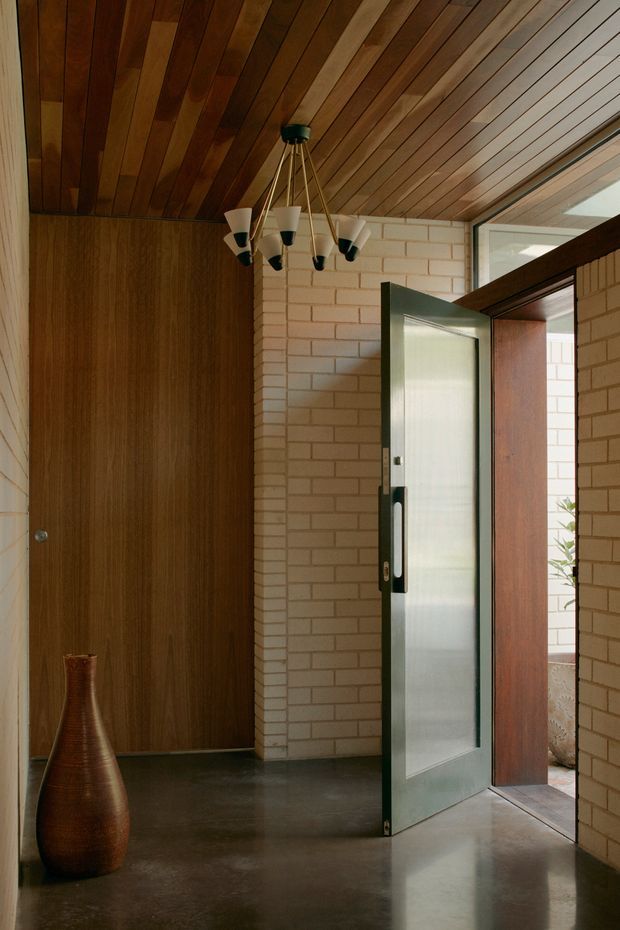 A mid-century-inspired entryway with a polished concrete floor, pale exposed brick walls, rich timber wall panelling and a timber-lined ceiling. A dark-framed frosted-glass door opens to an exterior courtyard, while a sculptural multi-arm pendant light and tall decorative vase add character.