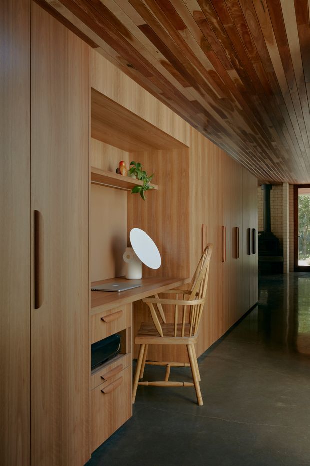A warm, narrow built-in home workspace and storage wall lined entirely in timber, with integrated cabinetry, open shelving, a desk, drawers, and a wooden chair. The timber-clad ceiling continues the material palette overhead. A small desk lamp, plant, and decorative objects sit on the shelves, while a polished dark floor and a corridor leading toward glazed doors extend into the background.