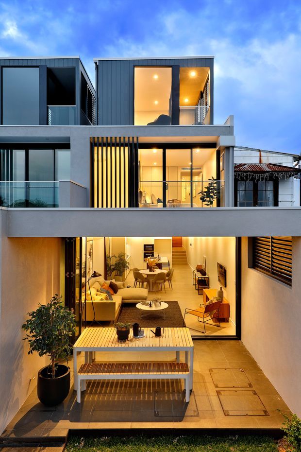 A dusk view of a contemporary multi-level urban residence with a deep central courtyard and open-plan living spaces. The façade combines dark vertical cladding, pale rendered walls, extensive black-framed glazing, glass balustrades, vertical sun-shading battens, and a partially covered rooftop or upper terrace. Warm interior lighting reveals a living room, dining area and stairwell, while the courtyard includes timber decking, built-in bench seating and potted planting.