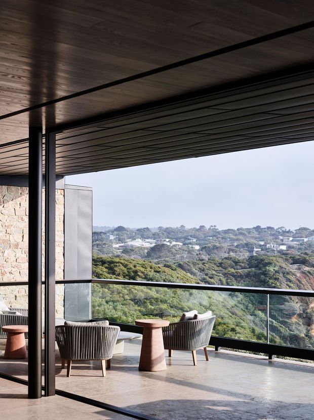 A covered outdoor terrace or balcony with expansive glazed balustrades overlooking a densely vegetated hillside. The space features dark timber-look soffit lining, black slender steel columns and framing, a stone-clad wall, polished concrete or large-format stone flooring, and contemporary outdoor lounge chairs with small sculptural side tables. The open-sided design and panoramic view create a refined, airy architectural setting.