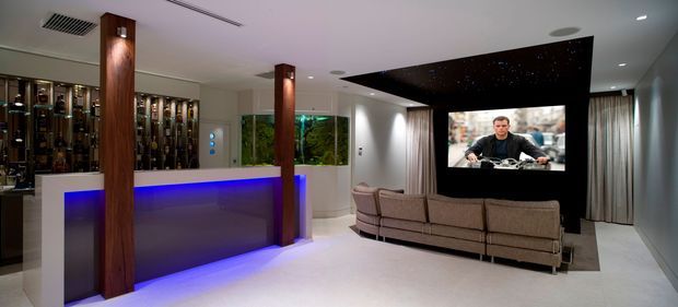 A contemporary home media and entertainment room with a large sectional sofa facing a wall-mounted projection screen or television. A sleek illuminated bar or display counter with blue LED underlighting occupies the left side, flanked by dark timber-clad columns. The room has a pale floor, recessed ceiling lighting, dark feature ceiling treatment above the screen, curtains, and built-in shelving displaying bottles or collectibles.