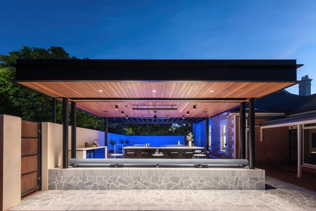 A contemporary covered outdoor entertaining area at dusk, featuring a broad flat-roof pavilion with a warm timber-lined ceiling, integrated lighting, dark structural framing, an outdoor kitchen and bar, and a stone-clad raised bench. Blue accent lighting illuminates the rear garden and adjacent brick house.