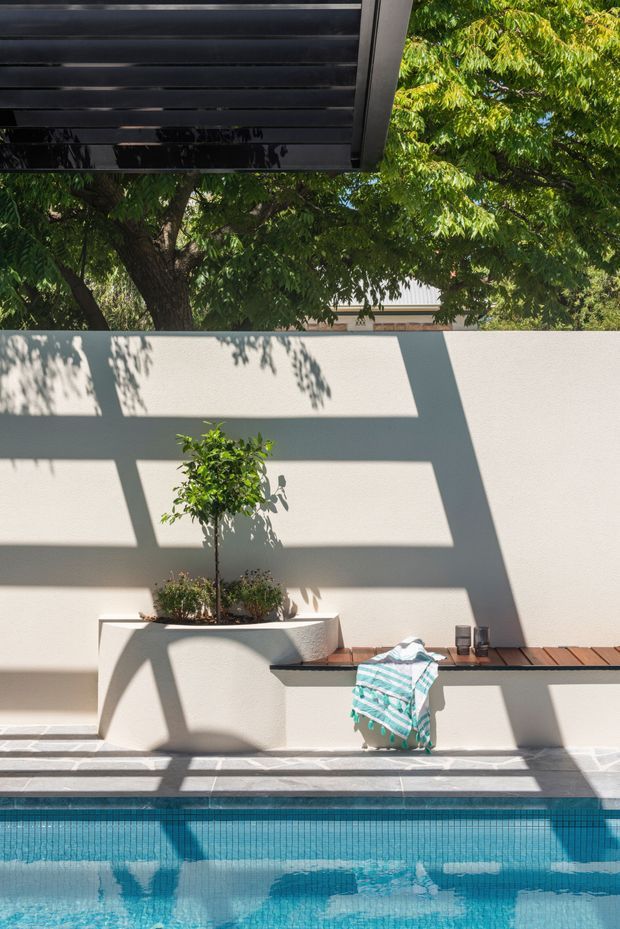 A bright outdoor poolside courtyard with a turquoise tiled swimming pool in the foreground, a white rendered boundary wall, integrated white planter, and a narrow timber bench or ledge. A small tree and planting cast strong geometric shadows across the wall and paving, while a dark overhead structure frames the upper left of the image. A folded striped towel and small objects rest on the bench, creating a relaxed, sunlit atmosphere.