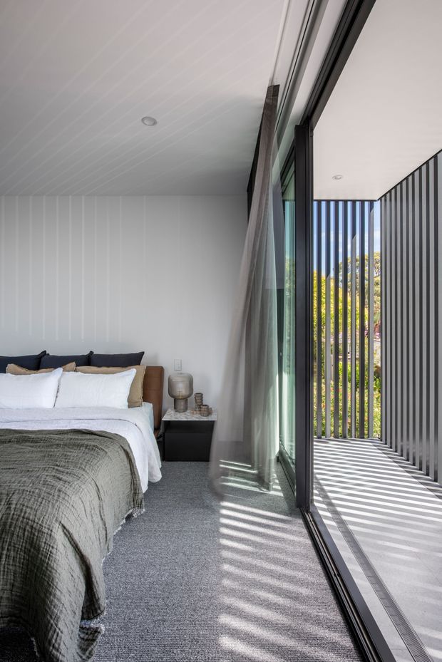 A bright contemporary bedroom opening onto a covered outdoor walkway or balcony through full-height glazed sliding doors. The room features a low bed with white bedding and a textured olive-grey throw, dark upholstered pillows, a warm brown bedhead, a black bedside table, grey carpet, and white vertically panelled walls and ceiling. Strong sunlight passes through the exterior vertical slatted screen, creating striped shadows across the carpet and pale outdoor paving.