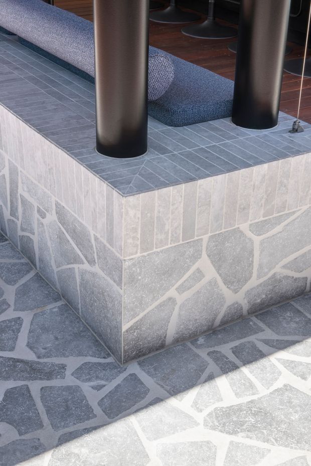 Close-up architectural detail of a raised terrace or entry platform clad in grey stone. The vertical face combines rectangular and irregular natural-stone pieces with pale grout, while the top surface is finished in small rectangular grey tiles. Two large dark cylindrical structural posts support an overhead element, and a blue upholstered bench is partially visible behind them.