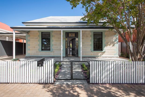 Street-facing view of a compact single-storey heritage-style residence with pale stone masonry, a corrugated metal hipped roof, dark-framed windows, and a small covered front entry porch. A low white vertical-slat fence with a central metal pedestrian gate encloses the landscaped front garden, while mature trees provide shade. The image is a bright architectural exterior photograph.