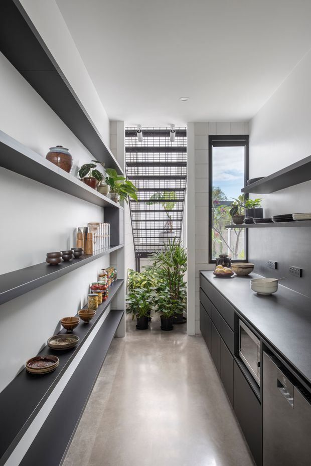A narrow, contemporary kitchen or scullery with full-height dark grey cabinetry and open display shelving along both sides. The space features a long dark benchtop, integrated appliances, polished concrete flooring, white walls, black-framed glazing, and a bright screened opening filled with indoor plants at the end. Ceramic bowls, jars, and potted greenery add natural accents.