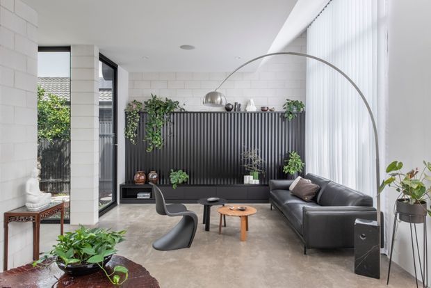 Bright contemporary living room with a polished concrete floor, white painted brick walls, full-height vertical blinds, and large glazing. A black ribbed feature wall incorporates low built-in joinery and open shelving with plants and objects. The room contains a black leather sofa, sculptural lounge chair, round timber coffee table, tall arched floor lamp, and indoor plants. The palette is predominantly white, black, grey, and warm timber, creating a refined yet inviting atmosphere.