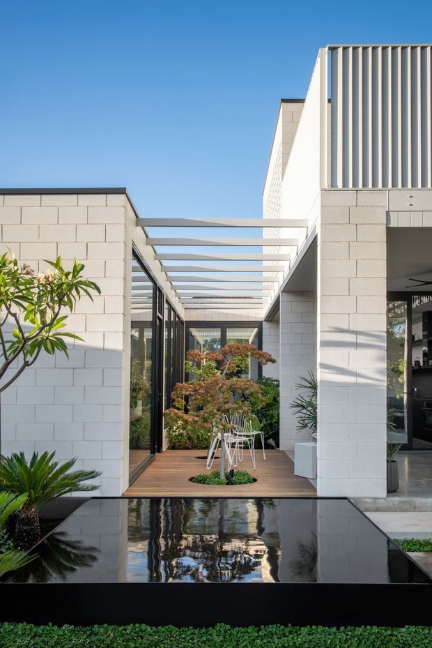 A contemporary courtyard-style residential exterior with pale masonry block walls, black-framed glazing, and a timber deck beneath a linear pergola. A shallow reflecting pool occupies the foreground, mirroring the architecture and planting. The composition includes strong geometric forms, vertical fins, lush courtyard landscaping, and a clear blue sky.