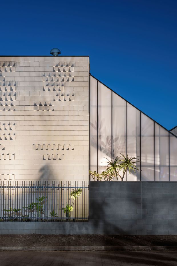 Exterior architectural view of a contemporary building under a clear deep-blue sky. The facade combines a pale masonry wall with patterned projecting brick details and a translucent, steeply sloping glazed or polycarbonate volume. A dark concrete-block boundary wall, metal security fencing, and planted areas occupy the foreground.