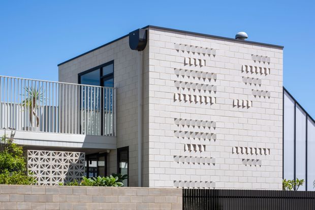 Exterior view of a contemporary two-storey building under a clear blue sky. The facade is composed of pale masonry blockwork with decorative geometric breeze-block openings arranged in patterned bands. Dark metal roof edging, window frames, balcony railings, and a black lower fence create contrast. A partially visible balcony, recessed entry, and planted areas are included in the foreground.