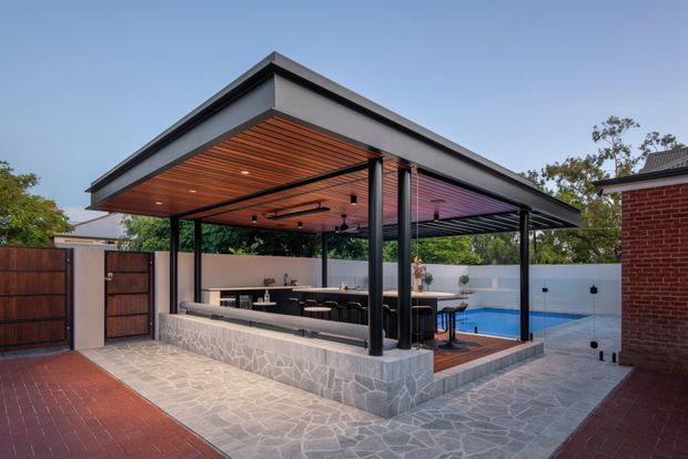 A contemporary covered outdoor entertaining pavilion beside a swimming pool, with a broad flat roof, dark metal posts and framing, a warm timber-lined ceiling, built-in outdoor kitchen or bar cabinetry, and extensive open sides. The pavilion sits on a paved courtyard with a low stone-clad perimeter wall and is adjacent to a brick residential building.