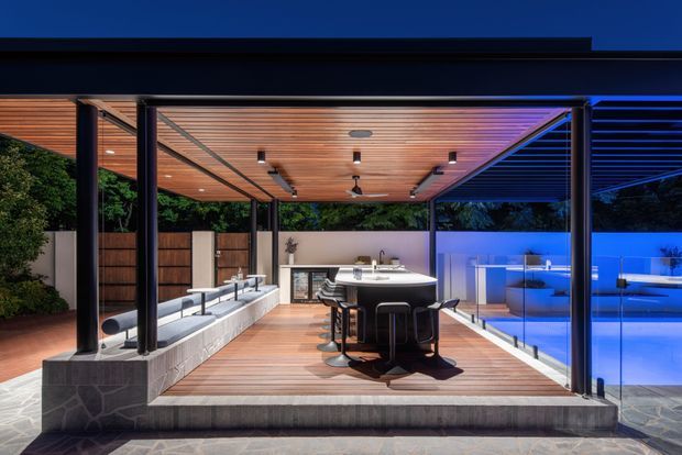 A sophisticated covered outdoor entertaining area beside a swimming pool, featuring a timber-decked dining zone with a long dark table and bar stools, a warm timber-lined ceiling, slender dark structural posts, glass pool fencing, and integrated outdoor lighting. The space opens onto landscaped greenery and is shown in dramatic evening blue lighting.