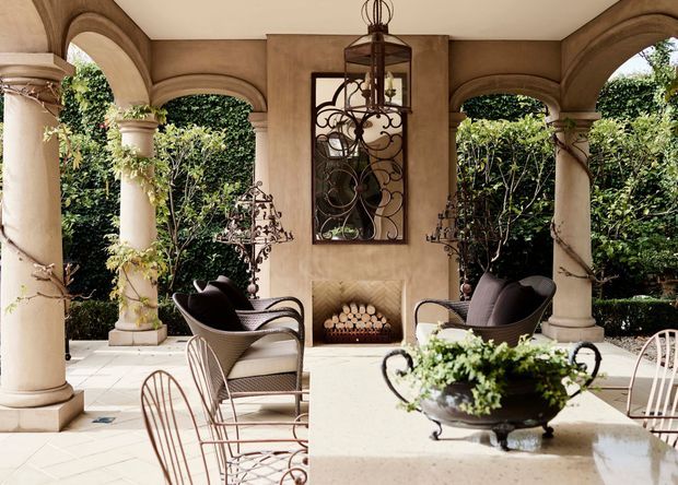 A refined covered outdoor terrace or loggia with arched openings, classical columns, a central fireplace, ornamental ironwork, and garden views. Wrought-iron seating and dining furniture surround a pale stone or concrete table with a floral planter. The space has a warm Mediterranean-inspired character with formal landscaping and decorative architectural detailing.