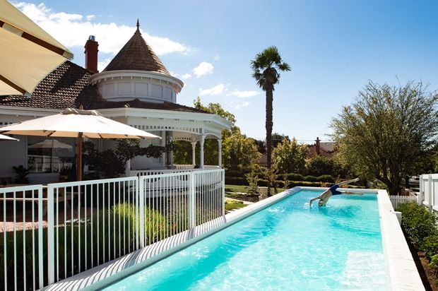 Exterior view of a grand heritage-style residence with a distinctive conical turret roof, ornate white veranda and balustrade, tiled roof, landscaped garden, and a long turquoise swimming pool in bright sunlight.