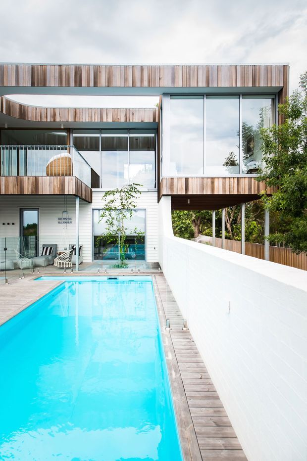 A contemporary multi-level home organized around a narrow outdoor swimming pool and deck. The facade combines extensive floor-to-ceiling glazing, vertical timber cladding, white rendered or painted masonry walls, and projecting box-like volumes. A glazed upper-level bridge and balcony overlook the pool courtyard, while mature planting is visible beyond the boundary wall.