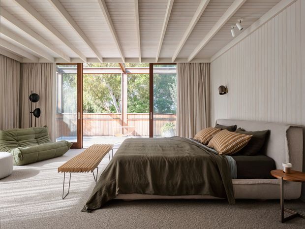 A warm, contemporary bedroom with a low upholstered bed, layered neutral and olive bedding, a ribbed timber coffee table, and a sculptural green lounge chair. Full-height glazed doors open to a timber balcony, while beige curtains, vertical wall panelling, and a white exposed-beam ceiling add texture and architectural character.