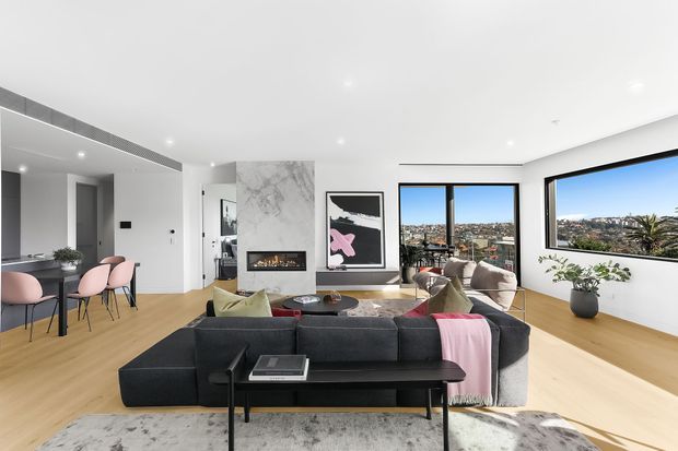 Bright contemporary open-plan living area with a dark sectional sofa, black coffee table, pale timber flooring, and a dining zone with blush-pink chairs. A marble-look fireplace feature wall, minimalist artwork, recessed lighting, and large corner windows overlook an urban landscape. The room has a clean, refined and airy feel.