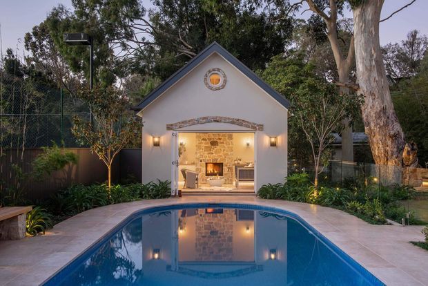 A landscaped backyard with a long rectangular swimming pool leading toward a small illuminated pool house or pavilion. The pavilion has a simple gabled facade, pale rendered walls, dark blue-grey trim, a round feature window, and wide open doors revealing a rustic stone fireplace wall and seating inside. Mature trees, planted garden beds, outdoor lighting, and paved pool surrounds create a warm evening setting.
