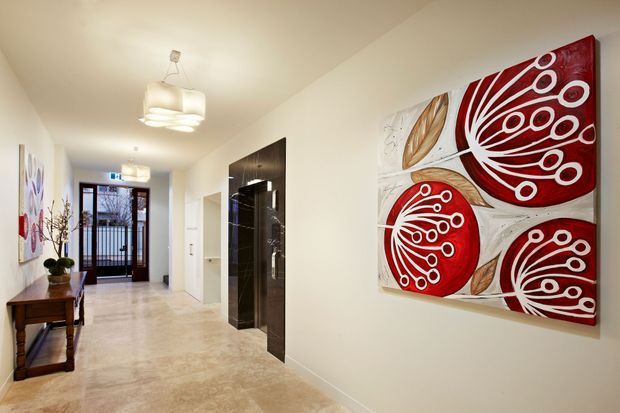 Bright contemporary residential hallway viewed toward a glazed entry door. The long corridor has pale walls, a polished light stone floor, modern pendant lights, dark timber or black cabinetry/door detailing, and a console table with decorative objects. Large bold red-and-white abstract floral artwork is mounted on the right wall, with additional artwork along the left wall.