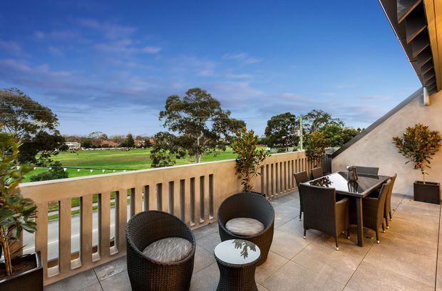 A spacious elevated outdoor terrace overlooking a manicured green landscape, with a concrete balustrade, tiled flooring, and dark woven outdoor lounge and dining furniture. A partial roof overhang and rendered wall frame the terrace, while potted plants add greenery. The scene is bright and open with expansive sky and distant trees beyond.