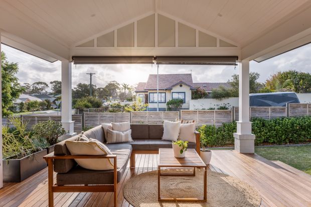 A bright, covered outdoor living area with a timber deck, built-in or modular sectional seating, armchairs, coffee table, rug and potted plant. The space has a high pitched gable roof with exposed decorative framing, white-painted posts and beams, retractable outdoor blinds, and views toward a suburban garden and neighbouring homes.