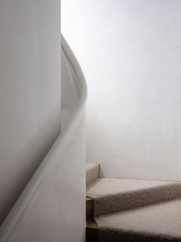 Minimalist interior view of a curved staircase turning around a central wall. The stair and surrounding walls are painted white, with pale textured carpet covering the treads and landing; a small portion of dark timber is visible at the stair edges. Soft natural light creates a calm, sculptural atmosphere.