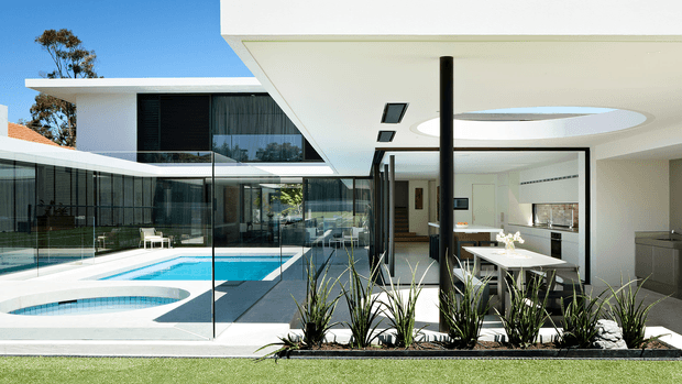 A bright contemporary residential courtyard viewed from the lawn, featuring a rectangular swimming pool, white geometric paving, extensive frameless-style glass fencing and glazing, and a flat-roofed white house. The covered outdoor living and dining area opens directly to the pool, with a large circular skylight, black structural column, minimalist dining table, and landscaped planting bed. The architecture uses crisp horizontal planes, floor-to-ceiling glazing, and strong indoor-outdoor connections.