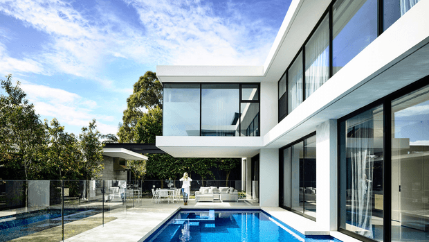A bright contemporary two-storey residence viewed from the pool courtyard. The white geometric form features extensive floor-to-ceiling glazing, projecting upper-level volumes, broad horizontal rooflines and a covered outdoor living area beside a rectangular swimming pool. Glass pool fencing, pale paving and landscaped trees surround the home.