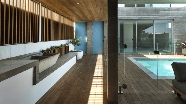 A modern indoor-outdoor transitional space with warm timber decking and ceiling boards, a tall vertical timber screen, white rendered planter walls, exposed concrete surfaces, and a glazed opening overlooking a turquoise swimming pool. Strong linear sunlight and reflections create a clean, architectural composition.