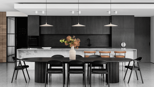 A sophisticated open-plan kitchen and dining area with a long dark kitchen wall, integrated cabinetry and appliances, a large pale stone island with three timber stools, and a substantial dark dining table surrounded by black chairs. Three pendant lights hang above the dining zone, while a vase of dried foliage provides a restrained natural accent. The space has a refined, monochromatic contemporary aesthetic with strong linear detailing and a polished floor.