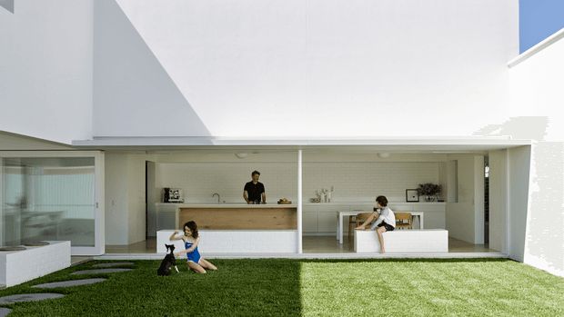 Bright contemporary courtyard interior-exterior composition viewed from the lawn, showing a white minimalist pavilion with large glazed openings, a linear kitchen and dining/work area beneath a broad overhang, and a carefully landscaped grass courtyard with stepping stones. The space has clean geometric lines, abundant daylight, and a double-height white wall volume.