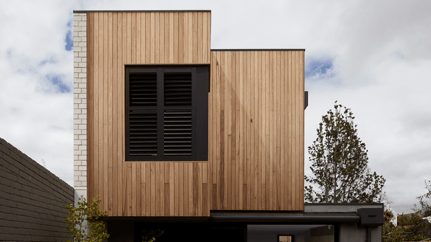 Exterior view of a contemporary multi-level building with two offset rectangular volumes clad in vertical light timber boards. A dark louvred window and dark flat roof edges contrast with the timber facade, while a pale masonry side wall and a partially visible lower level frame the composition. The image is photographed from outdoors under a cloudy sky.