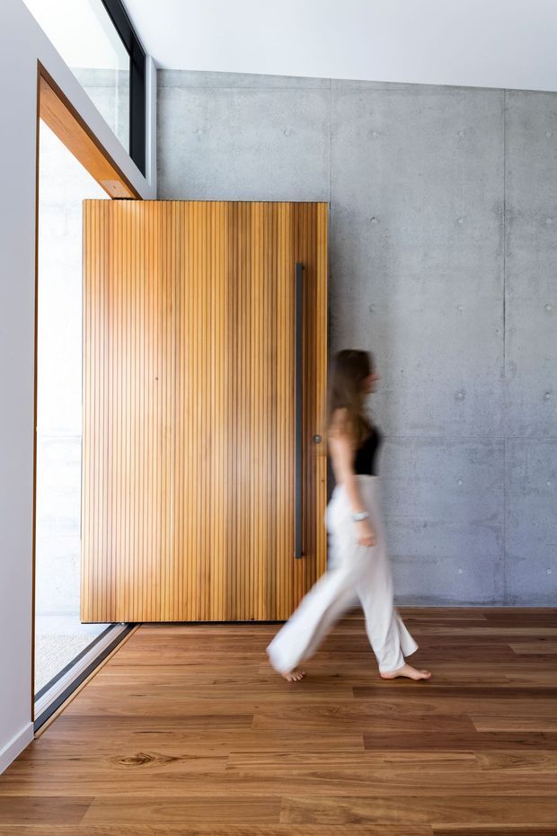 Modern interior featuring a tall pivot or entry door clad in vertical warm timber slats, set against a raw poured-concrete wall. The room has a timber floor, large black-framed glazing, and a high bright ceiling. A person walking through the space provides scale.