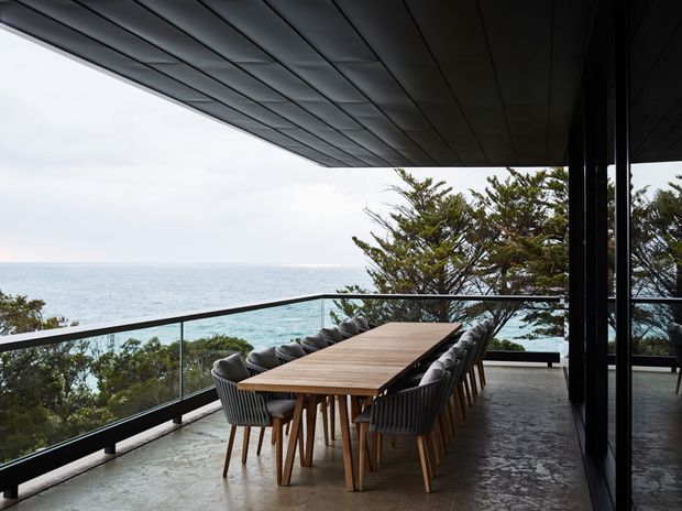 A covered outdoor dining terrace overlooking the ocean, with a long timber dining table surrounded by upholstered chairs. The terrace has a concrete floor, dark metal-framed floor-to-ceiling glazing, a glass balustrade, and a broad dark soffit, creating a contemporary coastal setting.