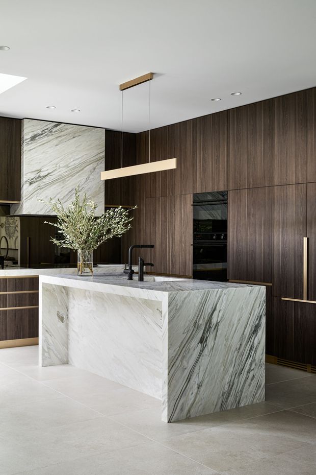 A refined contemporary kitchen featuring a large waterfall island clad in heavily veined white natural stone, dark timber veneer cabinetry spanning the rear wall, integrated black appliances, a black sink and tap, and a slim linear pendant light. Pale large-format floor tiles and a matching stone splashback create a high-contrast, luxurious interior.