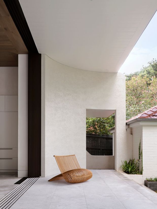 A contemporary covered courtyard or entry terrace with a broad white soffit, curved textured plaster wall, pale tiled paving, and a sculptural timber slatted lounge chair. Dark timber detailing frames a partially visible interior with built-in cabinetry on the left, while a small opening leads toward a planted boundary and an adjacent white brick outbuilding.