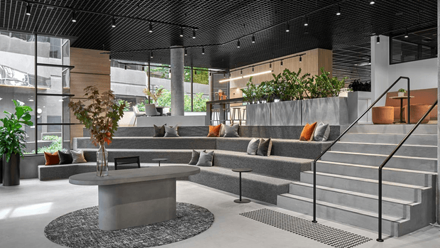 Contemporary multi-level commercial lounge or reception interior with broad tiered upholstered bench seating, low coffee tables, a central sculptural concrete table, indoor planting, and a stairway with black handrails. The space features extensive glazing, timber wall panelling, exposed concrete columns, and a black open-grid ceiling with track lighting.